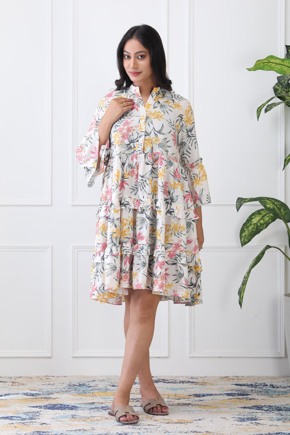 Woman wearing a floral dress standing in a room with a plant and white paneled wall.