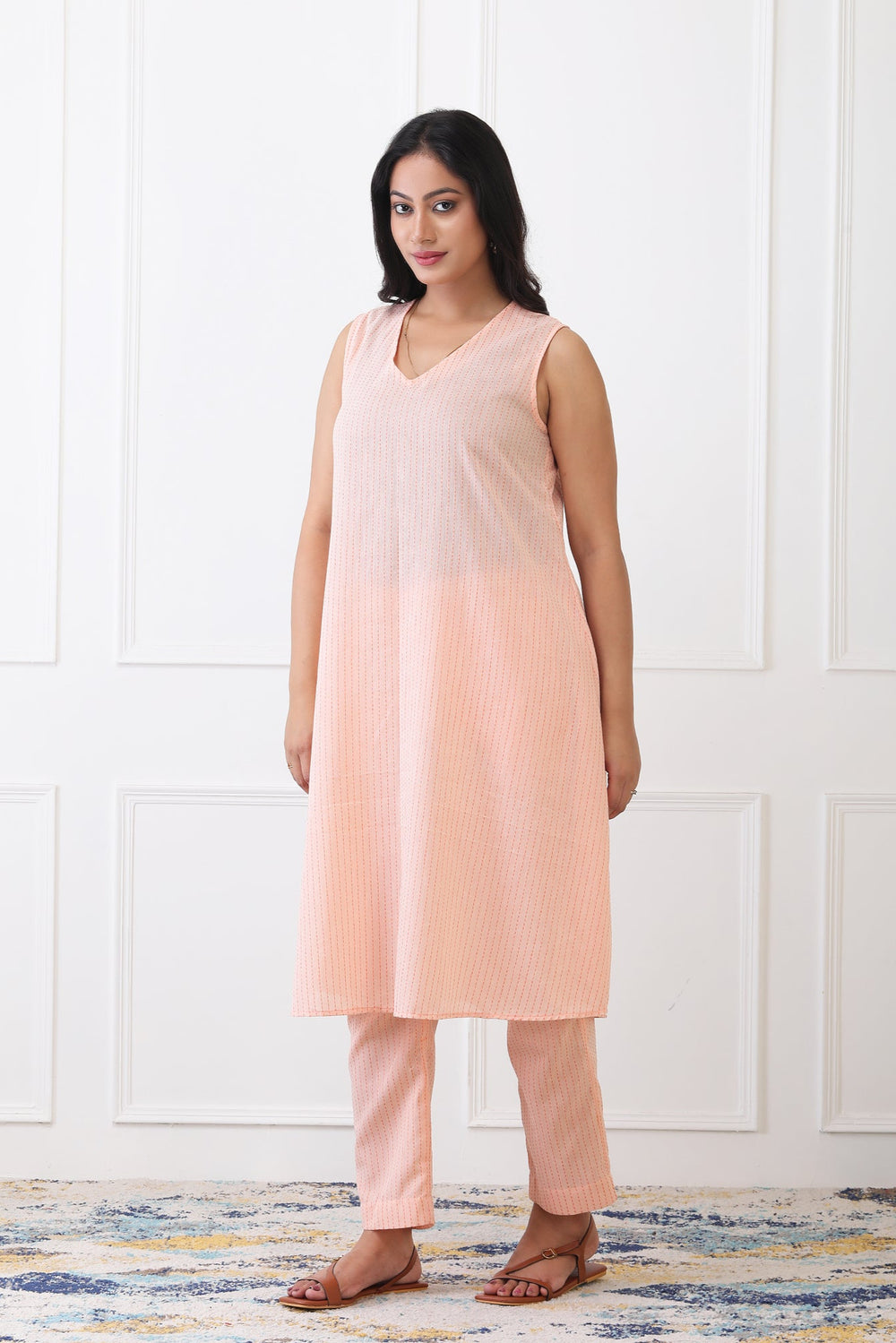 Woman wearing a peach sleeveless top and pants against a white paneled wall.