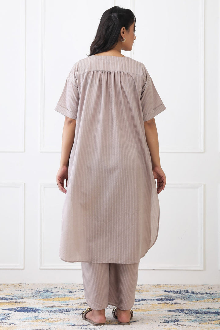 Woman wearing a beige striped dress and pants standing against a white paneled wall.