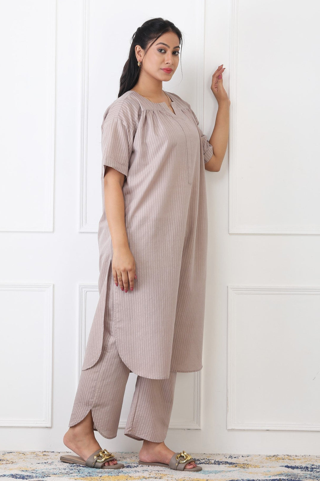 Woman wearing a beige outfit standing against a white paneled wall.