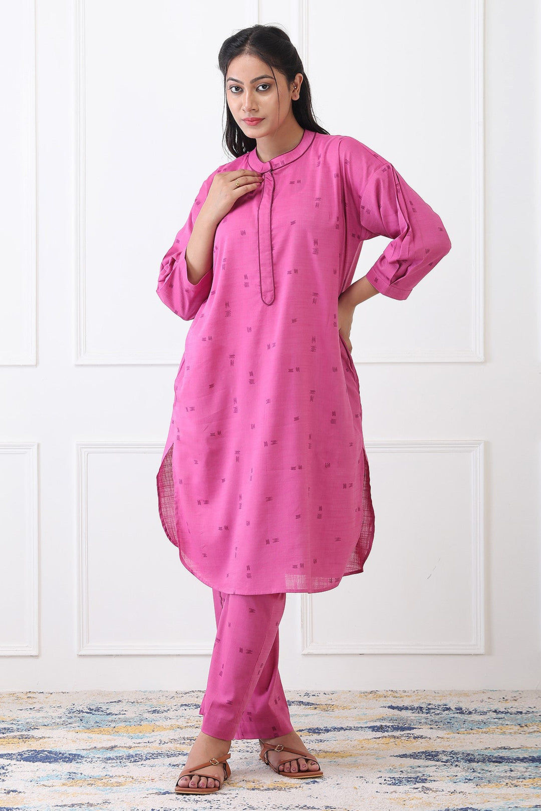 Woman wearing a pink kurta with matching pants against a white paneled wall.