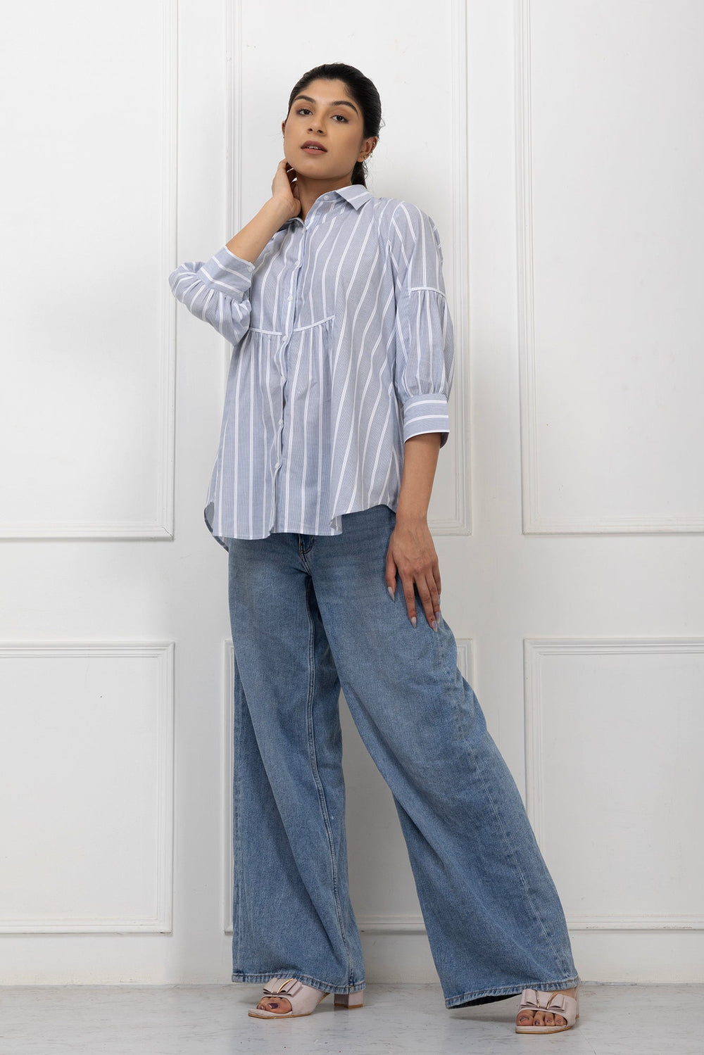 Woman wearing a striped shirt and wide-leg jeans against a white paneled wall.