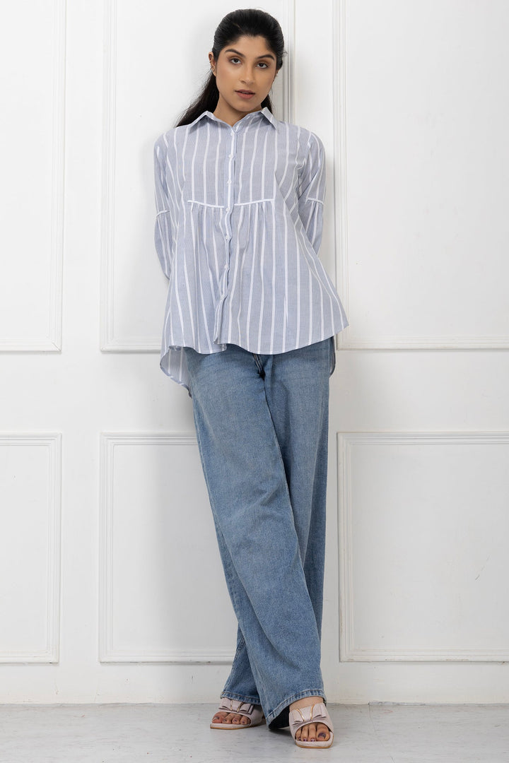 Woman wearing a striped shirt and jeans standing against a white paneled wall.