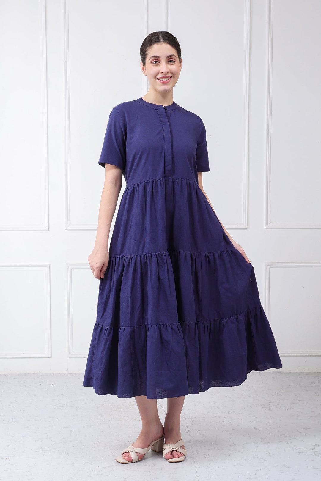 A woman wearing a short-sleeved, tiered midi dress in deep purple, standing against a white paneled wall.