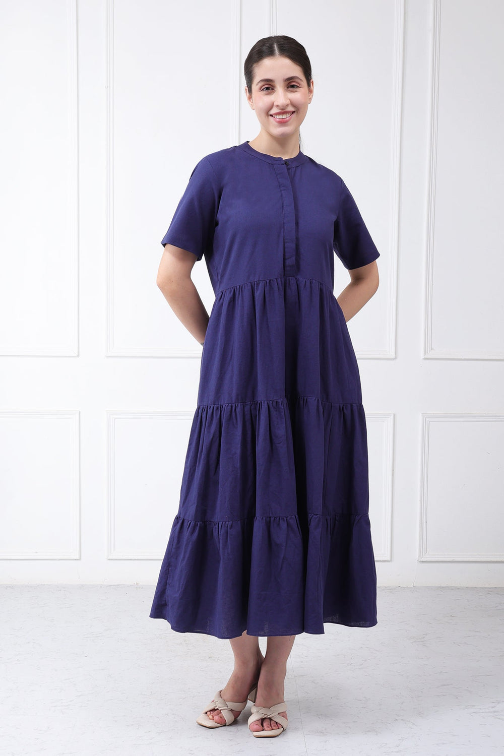 A woman modeling a short-sleeved, tiered midi dress in a deep purple color, standing against a white paneled wall.