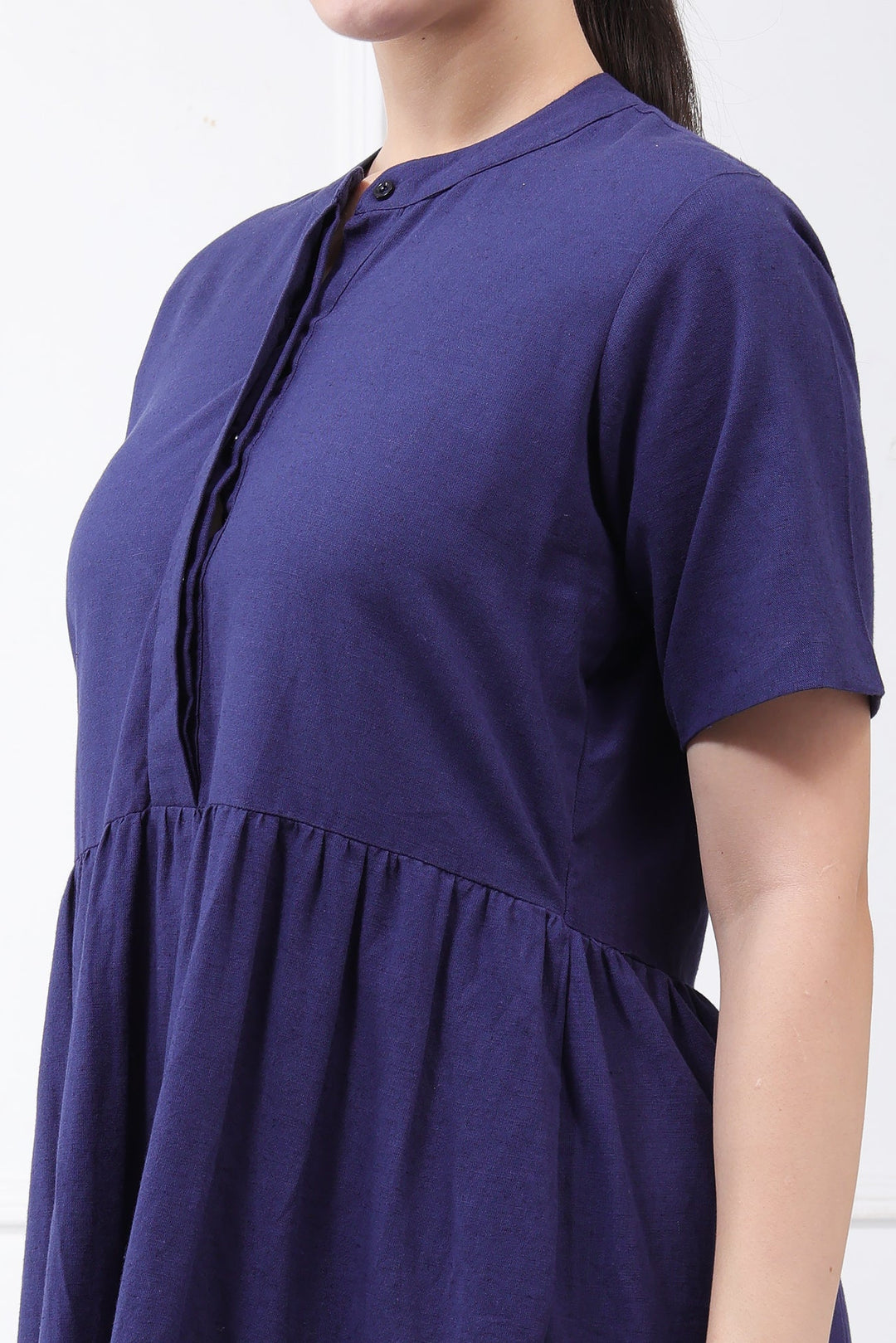 A close-up side view of a woman wearing a short-sleeve, high-neck dress in a deep indigo blue. The dress features a gathered waist and a buttoned front placket.