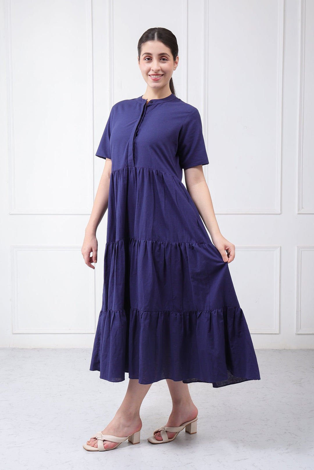 A woman modeling a short-sleeved, tiered midi dress in a deep purple shade, standing against a white paneled wall.