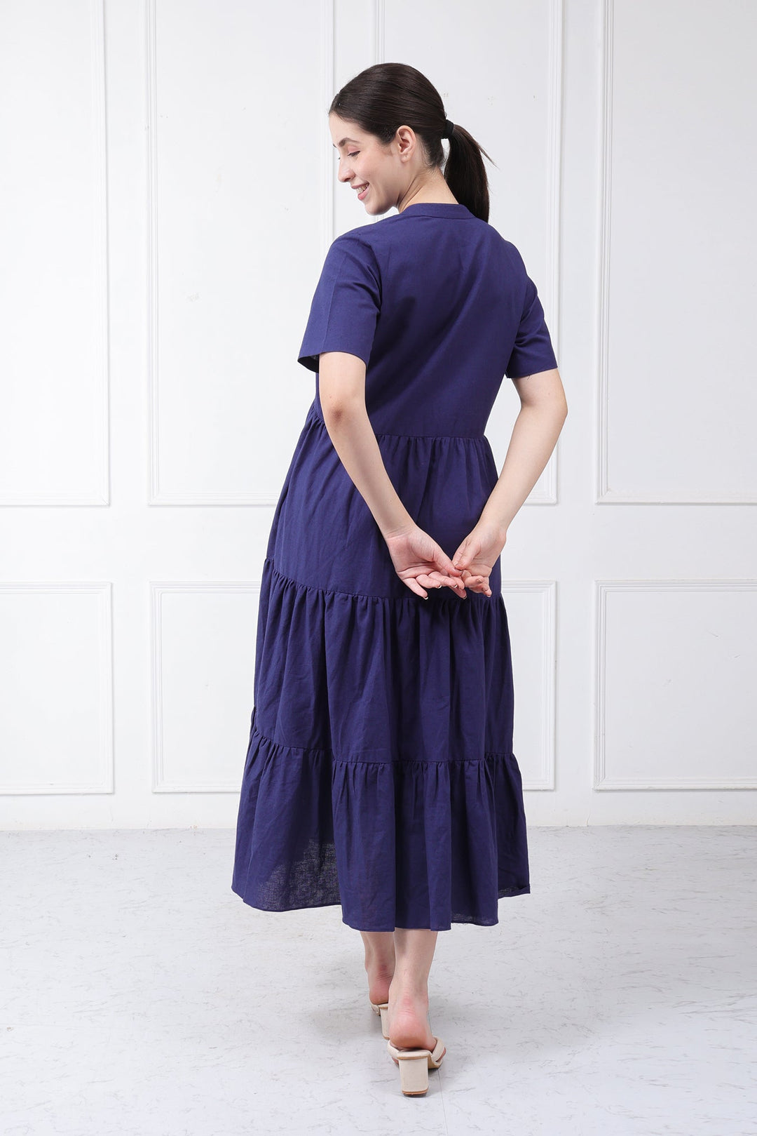 A woman wearing a short-sleeve, tiered midi dress in a deep purple shade, shown from the back against a white paneled wall.