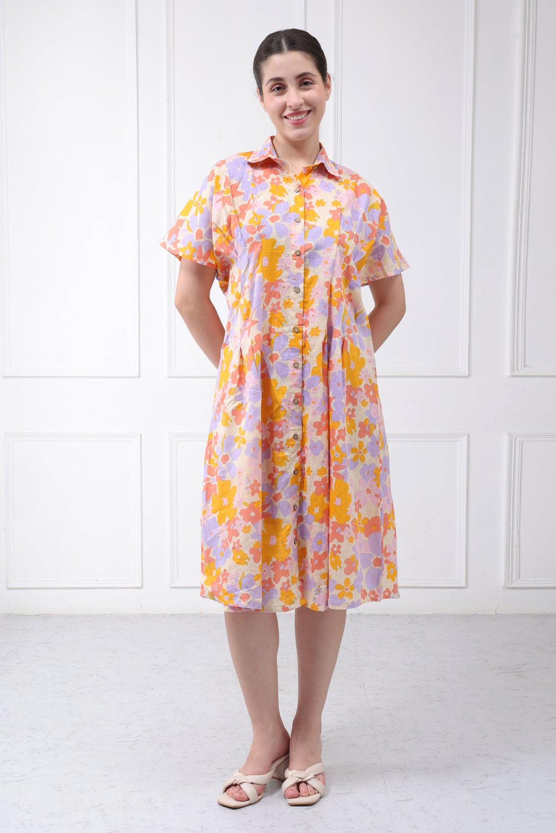 A woman modeling a short-sleeved button-up shirt dress with a vibrant orange, yellow, and purple floral pattern, standing against a white paneled wall.