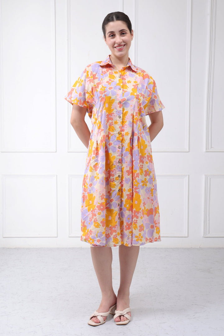 A woman modeling a short-sleeved button-up shirt dress with a vibrant orange, yellow, and purple floral pattern, standing against a white paneled wall.