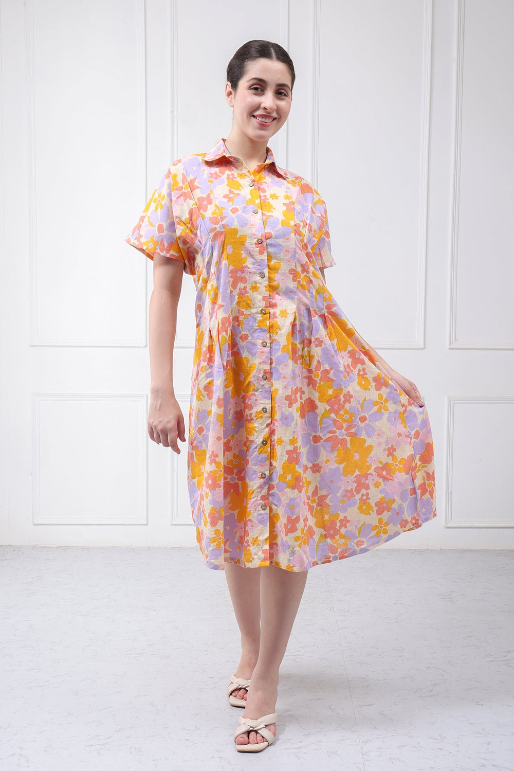 A woman modeling a short-sleeve button-up dress with a colorful floral pattern in orange, yellow, and purple on a light lavender base, set against a white paneled wall.