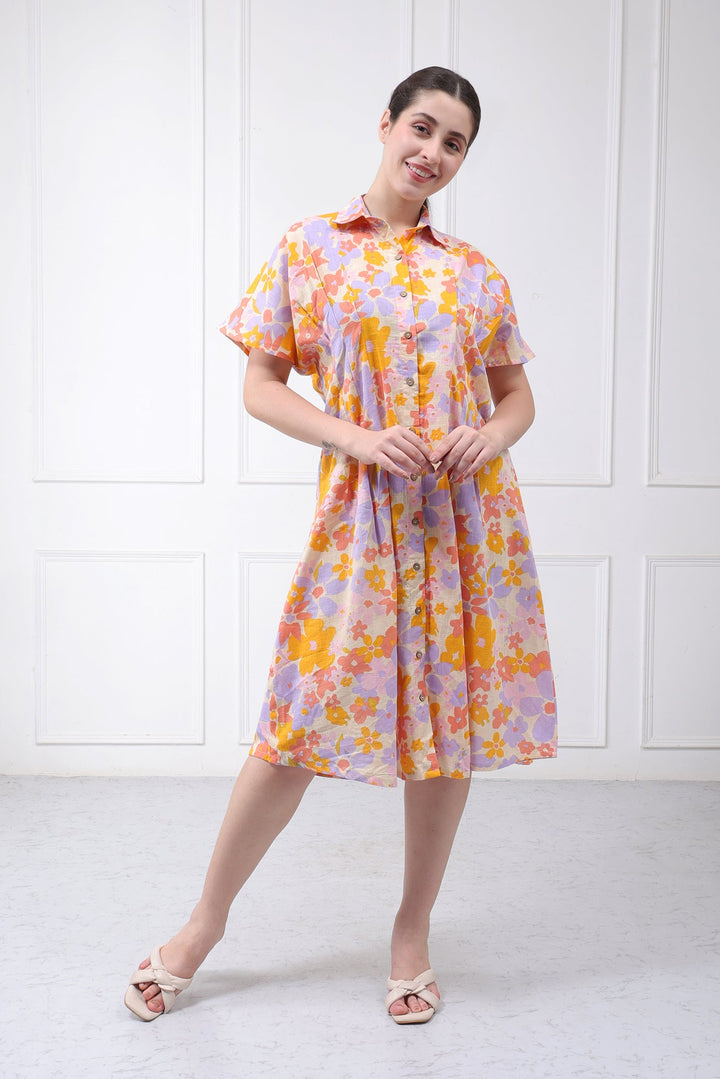 A woman modeling a short-sleeve button-up dress with a retro floral pattern in orange, yellow, and purple on a light pinkish-purple base, paired with light pink sandals, set against a white paneled wall.