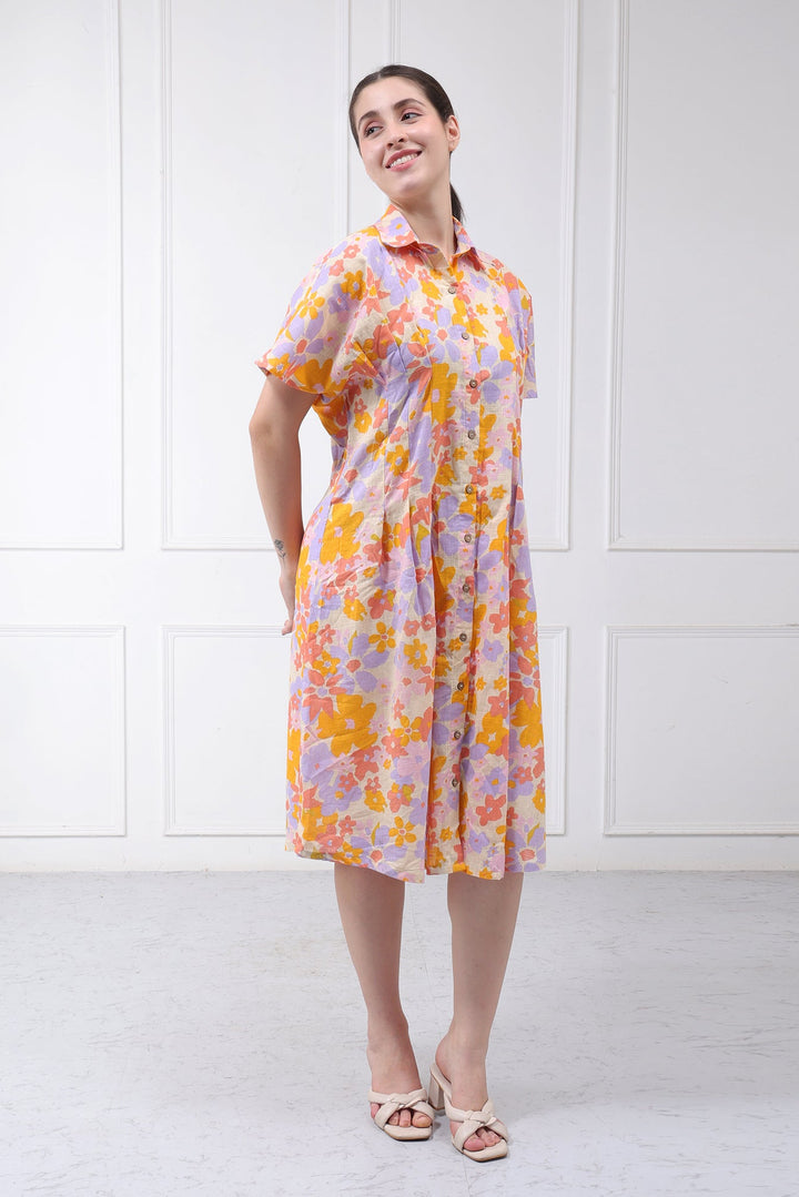 A woman modeling a short-sleeved, knee-length collared dress with a vibrant yellow, orange, and purple floral pattern on a light pinkish-purple base, set against a white paneled wall.