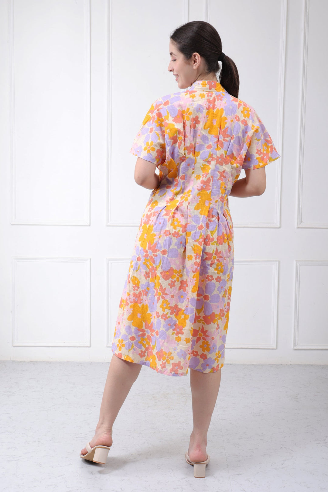 A woman wearing a short-sleeved floral midi dress with a purple and yellow pattern, photographed from behind against a white paneled wall.