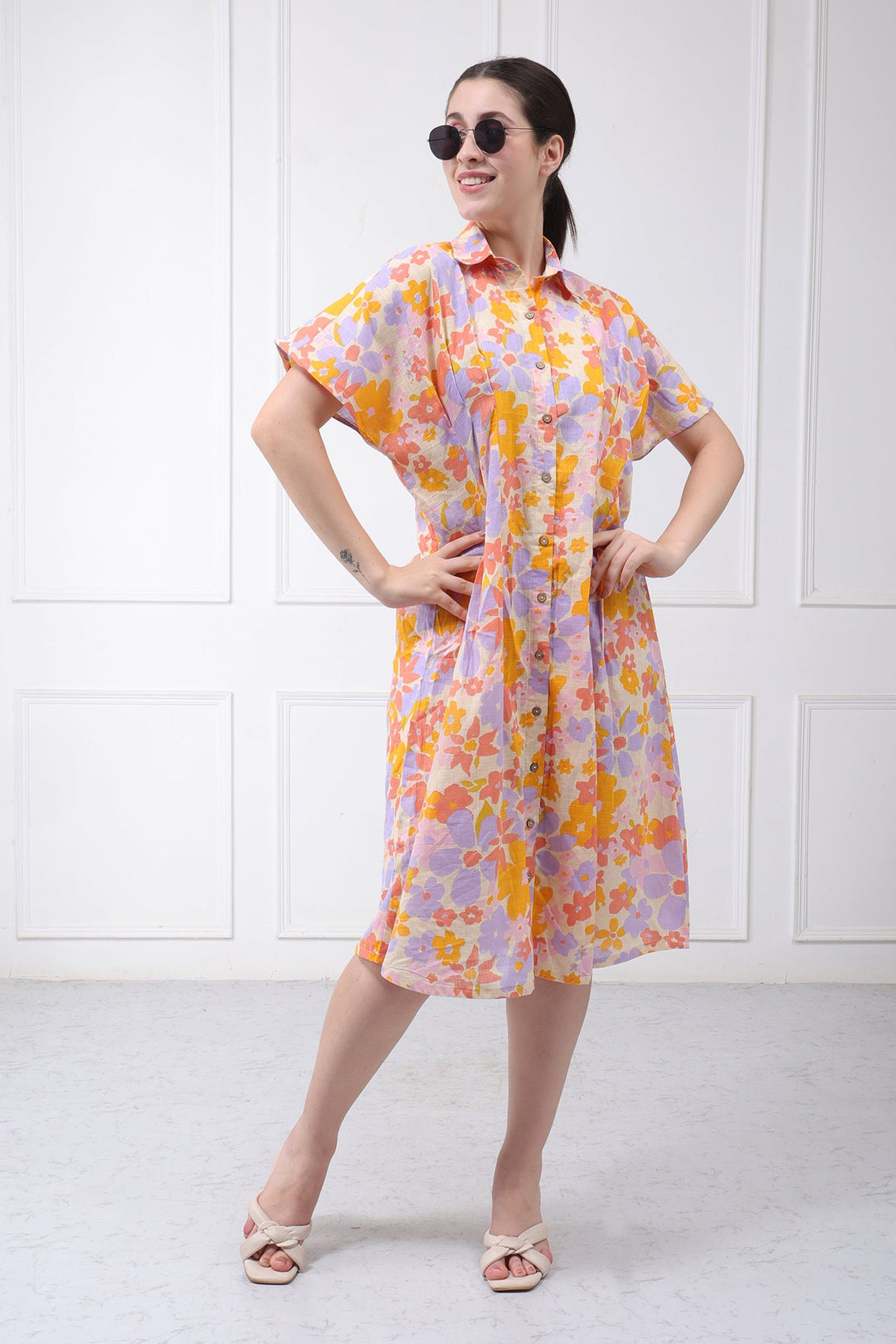 A woman modeling a short-sleeve collared dress with a vibrant orange, yellow, and purple floral pattern on a light pinkish-purple base, styled with sunglasses and beige sandals against a white paneled wall.