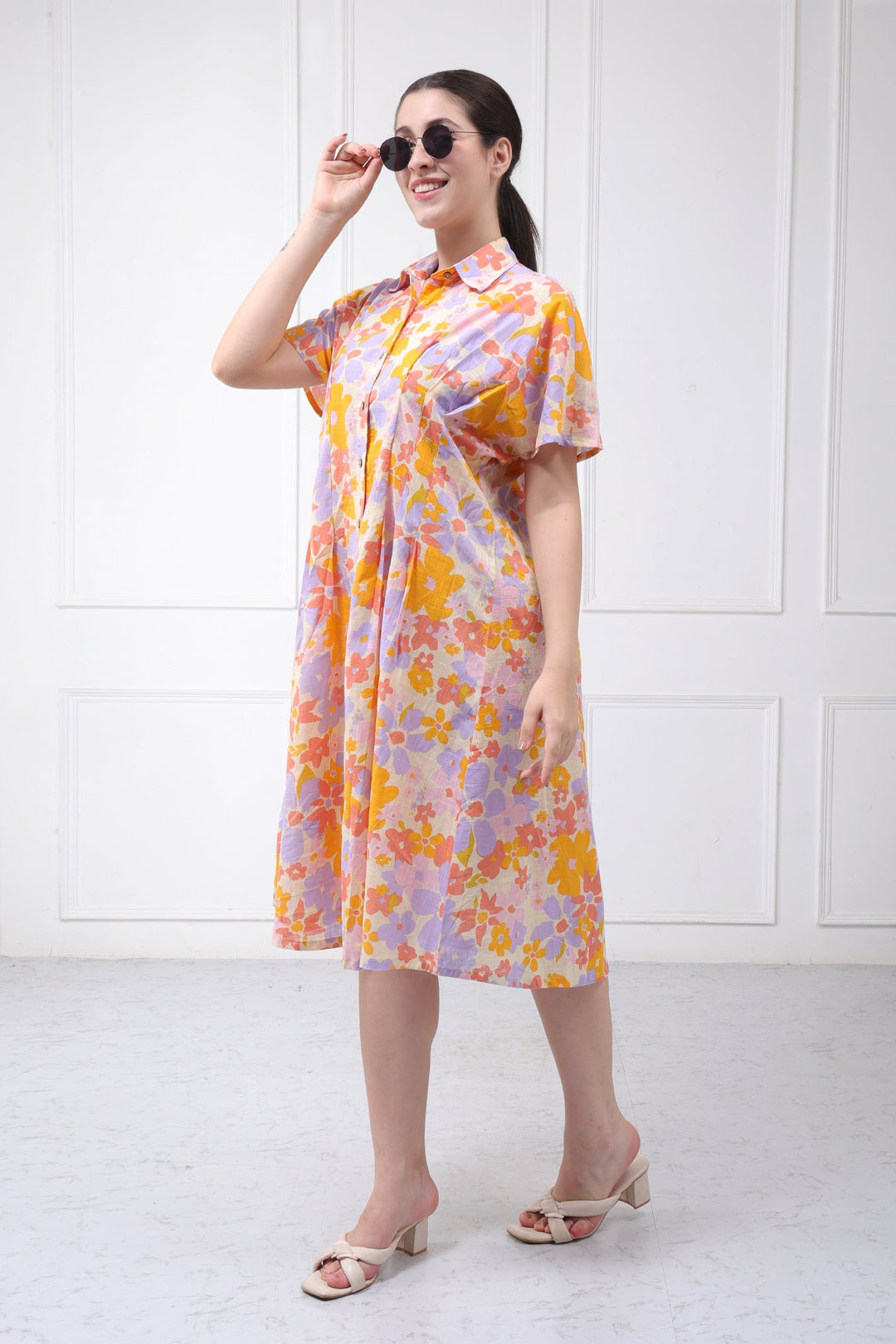A woman modeling a short-sleeve collared midi dress with a colorful floral print in orange, yellow, and purple on a light pinkish base, paired with beige heeled sandals and sunglasses, set against a white paneled wall.