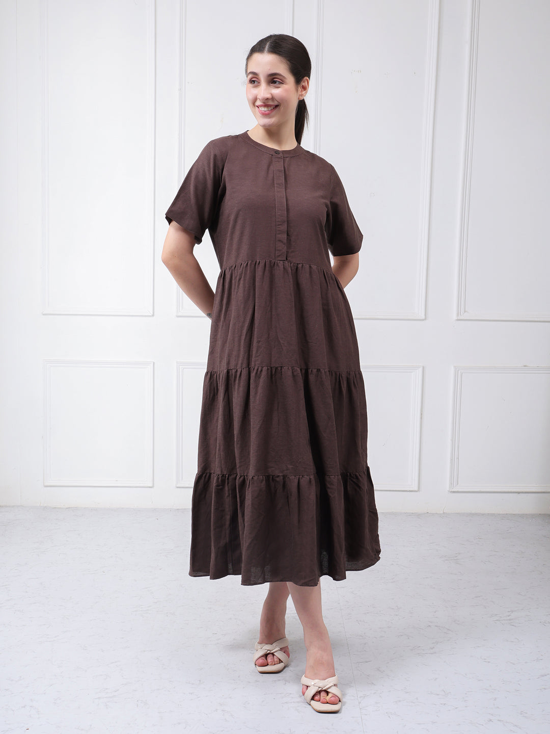 A woman modeling a dark brown tiered linen midi dress with short sleeves and a round neckline, standing against a white paneled wall.