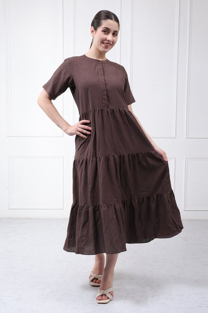 A woman modeling a dark brown tiered midi dress with short sleeves and a gathered waist, standing against a white paneled wall.