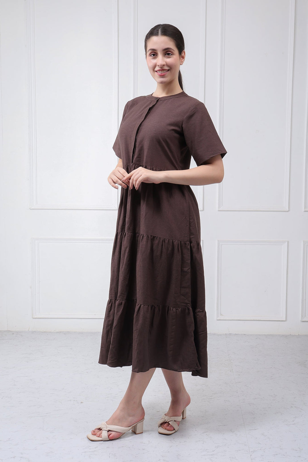 A woman modeling a short-sleeved, tiered midi dress in dark brown, standing against a white paneled wall.