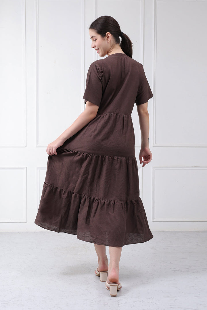 A woman wearing a dark brown tiered midi dress with short sleeves, shown from the back in a bright, white paneled room.