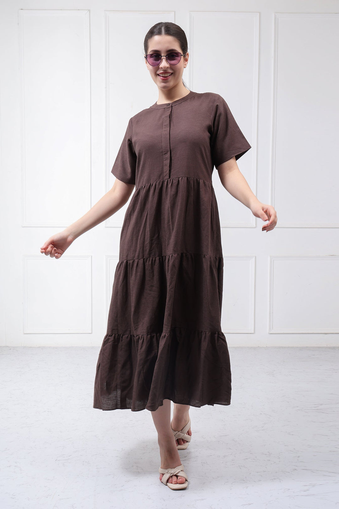 A woman wearing a dark brown short-sleeve tiered midi dress with a buttoned neckline, walking in front of a white paneled wall.