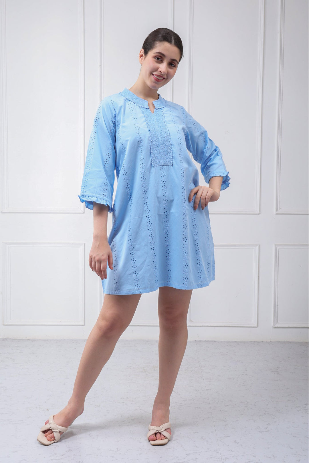 A woman modeling a light blue cotton kurta dress with three-quarter sleeves and a small keyhole cutout at the collar, standing against a white paneled wall.