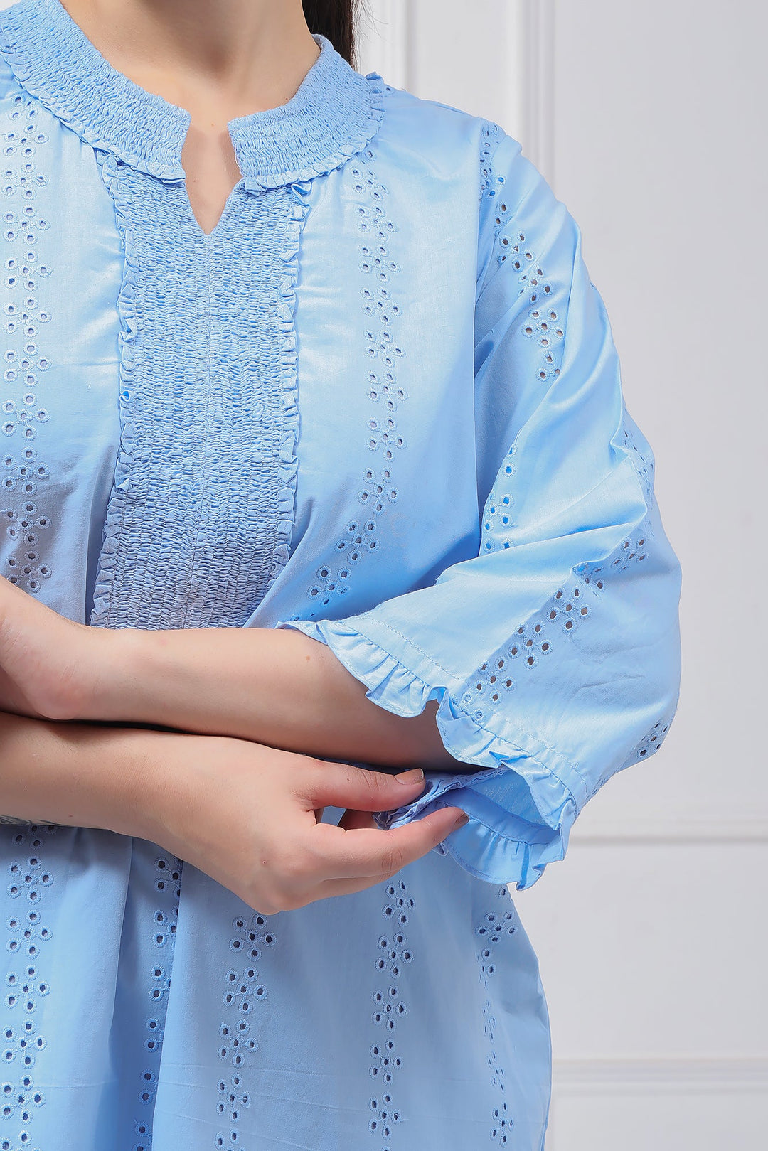 A close-up of a person wearing a light blue eyelet blouse with a split neckline, textured front panel, and ruffled elbow-length sleeves.