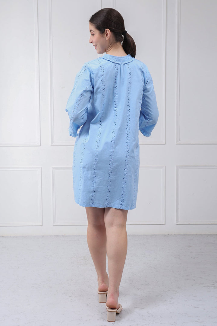 A woman viewed from behind wearing a light blue long-sleeve shirt dress with vertical eyelet embroidery, standing against a white paneled wall.