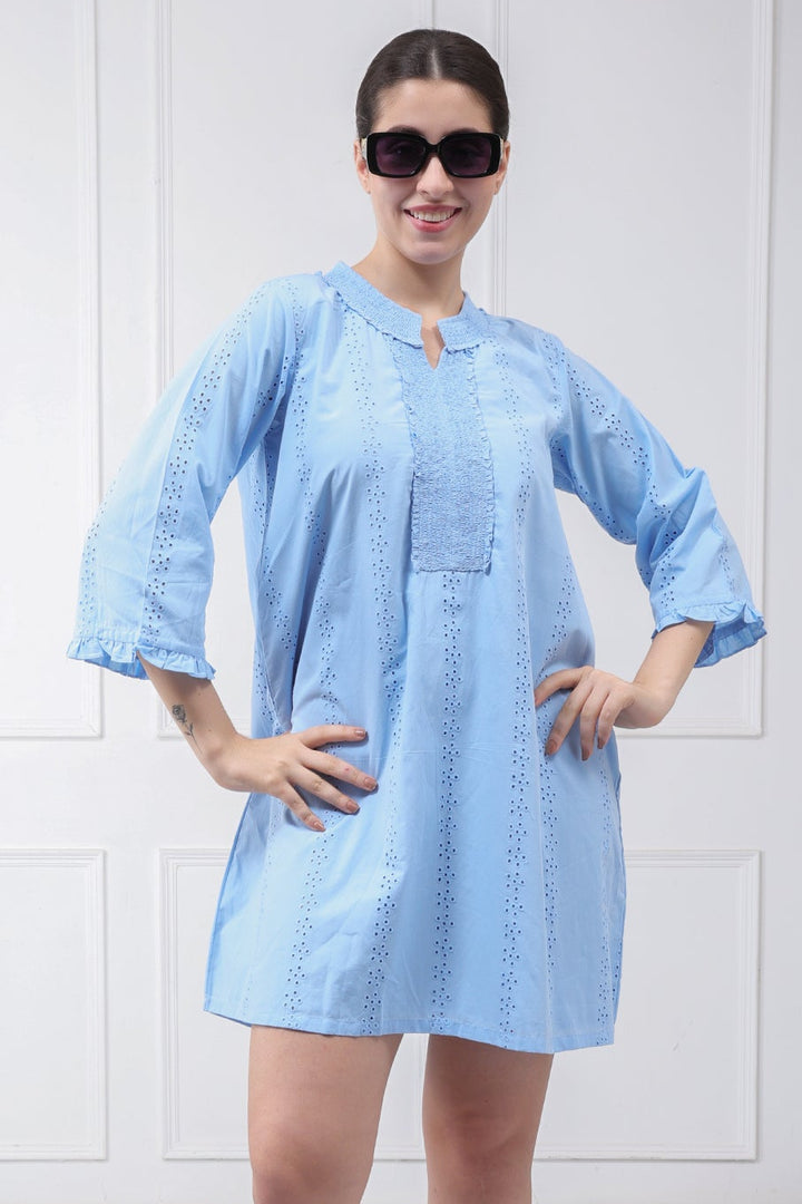 A woman modeling a light blue cotton tunic dress with embroidered details, paired with beige sandals and black sunglasses, standing against a white paneled wall.