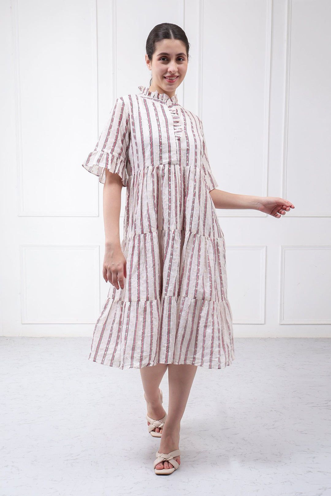 A woman modeling a tiered midi dress with vertical pink and white stripes, featuring a high mock neck, ruffled short sleeves, and a button-up front, set against a white paneled wall.