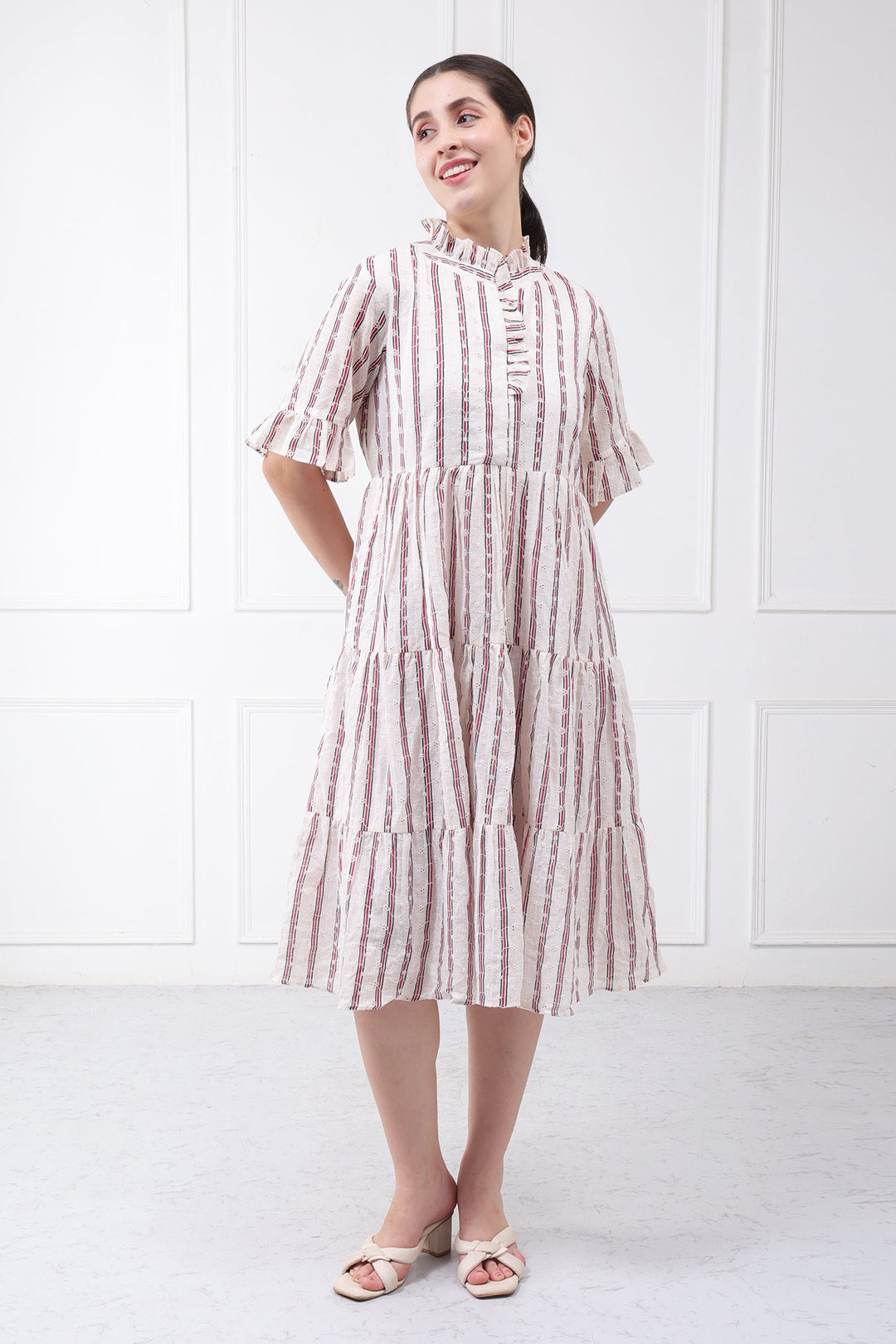 A woman modeling a tiered midi dress with vertical pink and white stripes, featuring a ruffled high neckline and elbow-length sleeves, paired with beige flat sandals against a white paneled wall background.