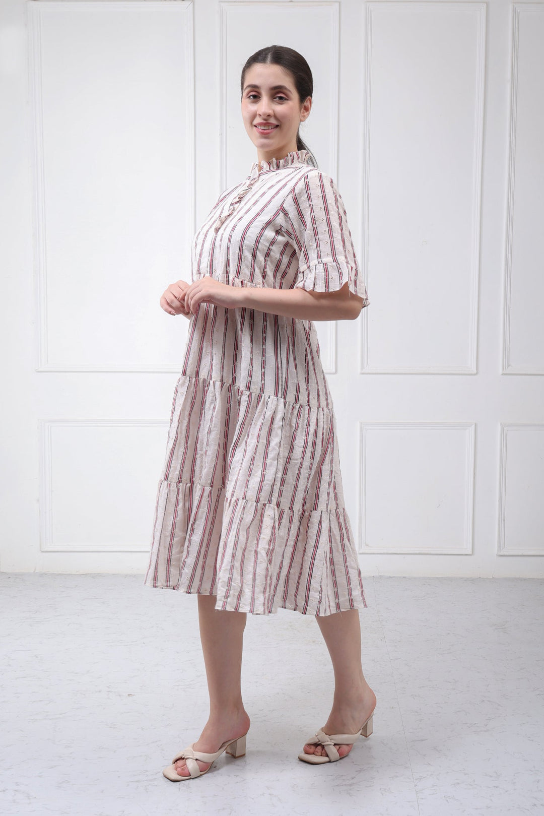 A woman modeling a tiered midi dress with vertical pink and beige stripes, standing in front of a white paneled wall.