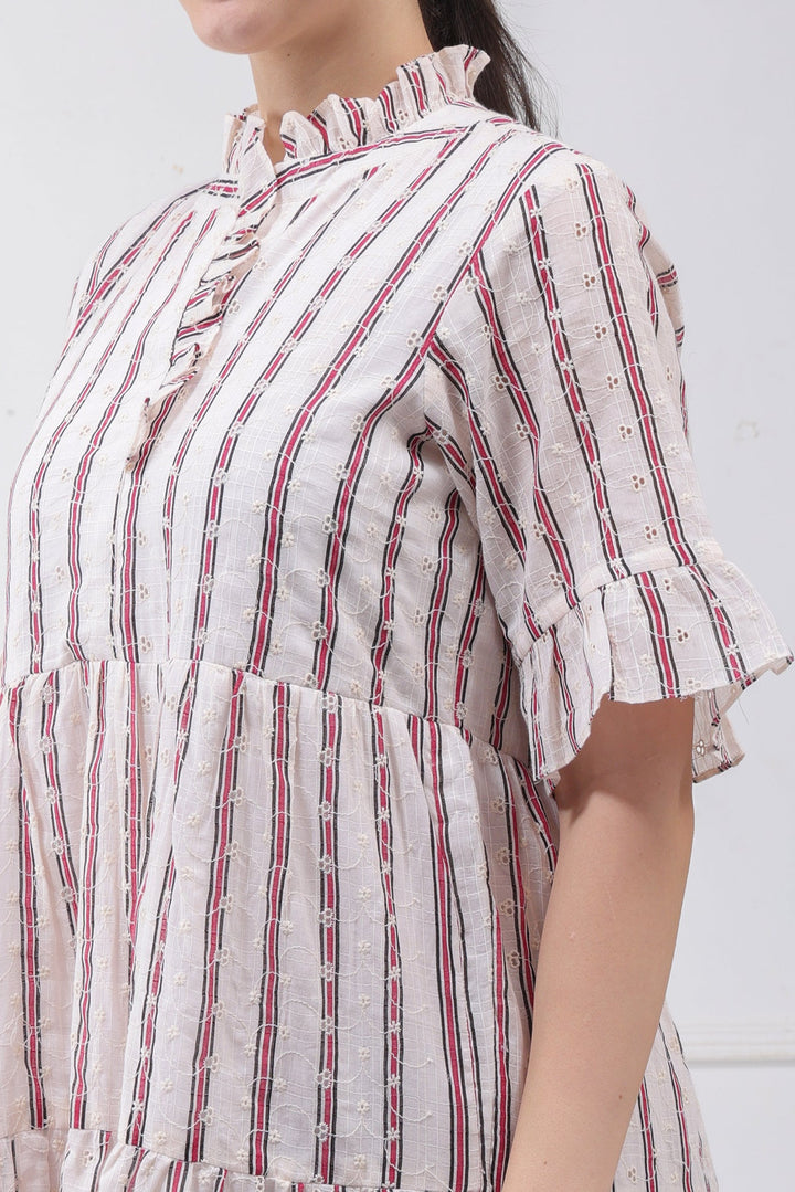 A close-up of a model wearing a short-sleeve blouse with vertical red and blue stripes on a white textured fabric, featuring a ruffled high collar and ruffled cuffs.