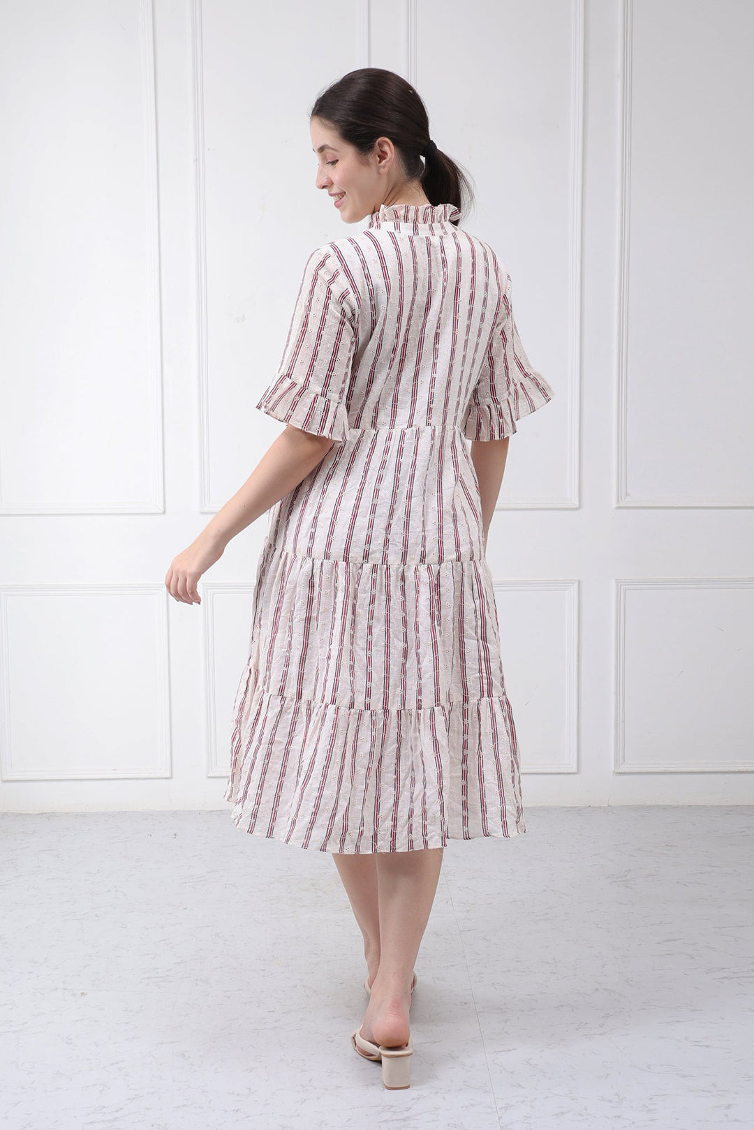 A woman wearing a tiered midi dress with vertical pink and white stripes, shown from the back against a white paneled wall.