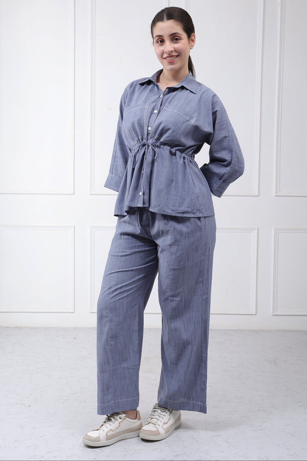 A woman modeling a matching two-piece lounge set in a dusty blue-gray color, featuring a long-sleeve button-up top with a tie waist and matching wide-leg pants, paired with white sneakers against a white paneled wall.
