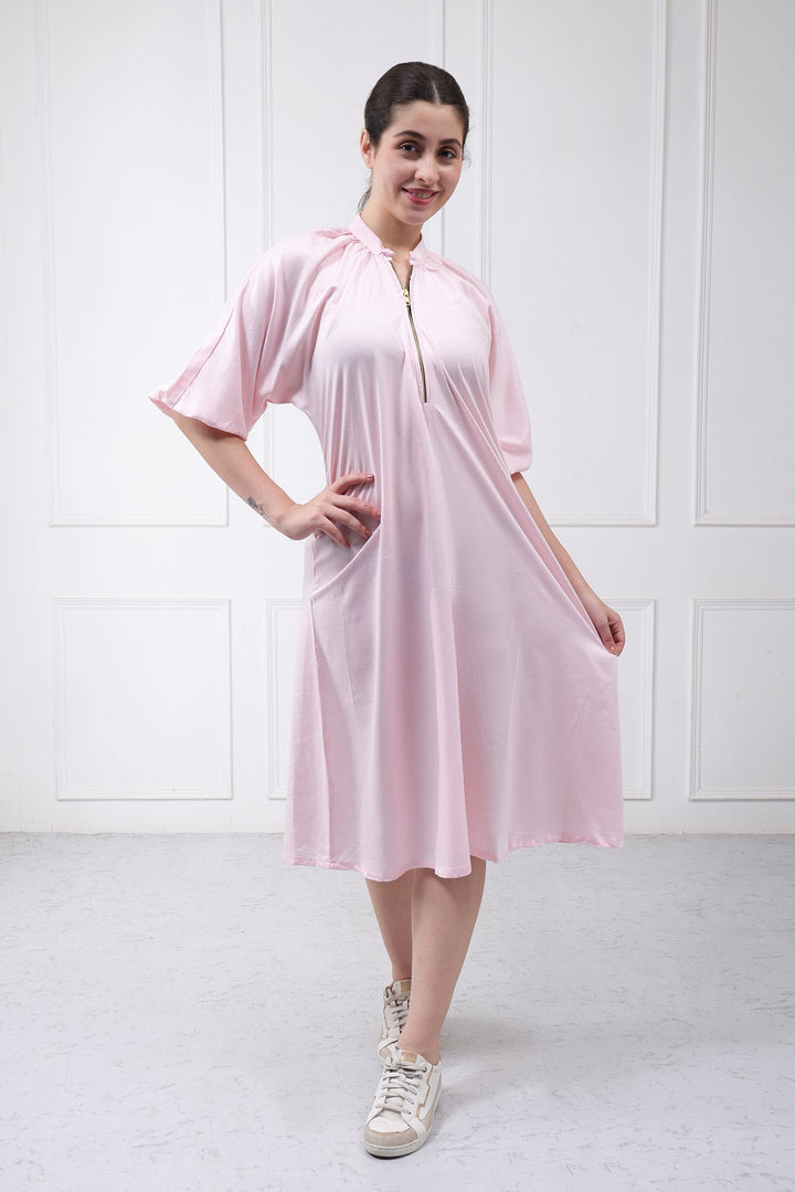 A woman modeling a light pink short-sleeve midi dress with a split neckline and front zipper, paired with white sneakers, standing against a white paneled wall.