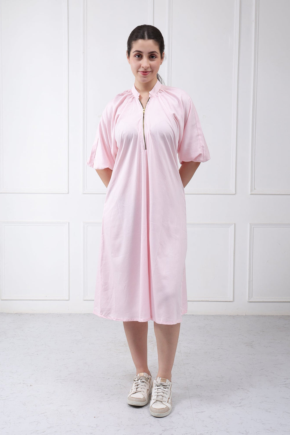 A woman wearing a light pink short-sleeve midi dress with a front zipper and drawstring tie, paired with beige sneakers, standing against a white paneled wall.
