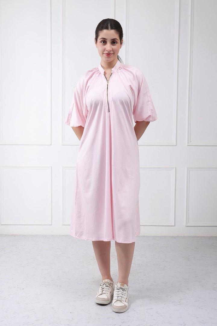 A woman wearing a light pink short-sleeve midi dress with a front zipper and drawstring tie, paired with beige sneakers, standing against a white paneled wall.