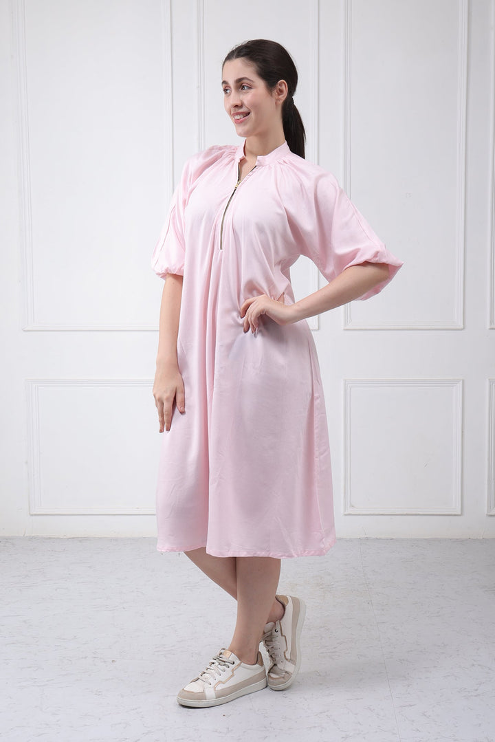 A woman modeling a light pink short-sleeve midi dress with a front zipper and stand collar, paired with beige sneakers, standing against a white paneled wall.
