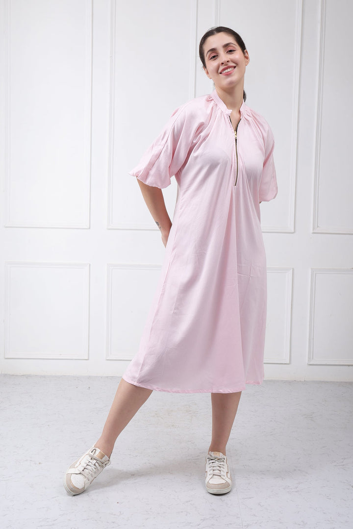 A woman modeling a light pink short-sleeve tunic dress with a front zipper, standing in a bright white room with paneled walls and light gray flooring.