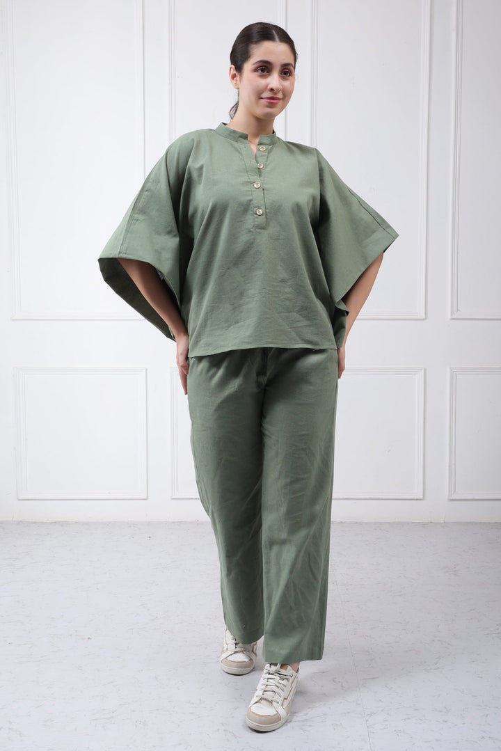 A woman modeling a matching sage green two-piece lounge set consisting of a short-sleeve button-up top and wide-leg trousers, paired with white sneakers, set against a white paneled wall.