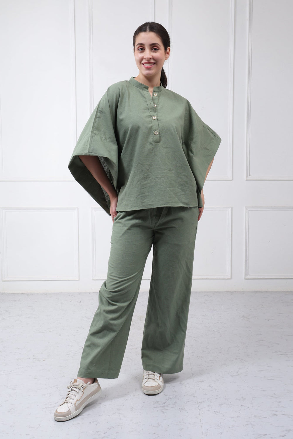 A woman modeling a matching sage green two-piece lounge set consisting of a loose, short-sleeved button-up top and wide-leg trousers, paired with white sneakers, set against a white paneled wall.