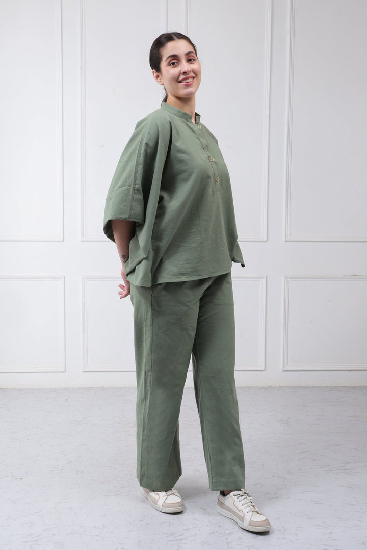 A woman modeling a matching sage green two-piece linen set consisting of a short-sleeve top with a mandarin collar and wide-leg trousers, paired with white sneakers, set against a white paneled wall.