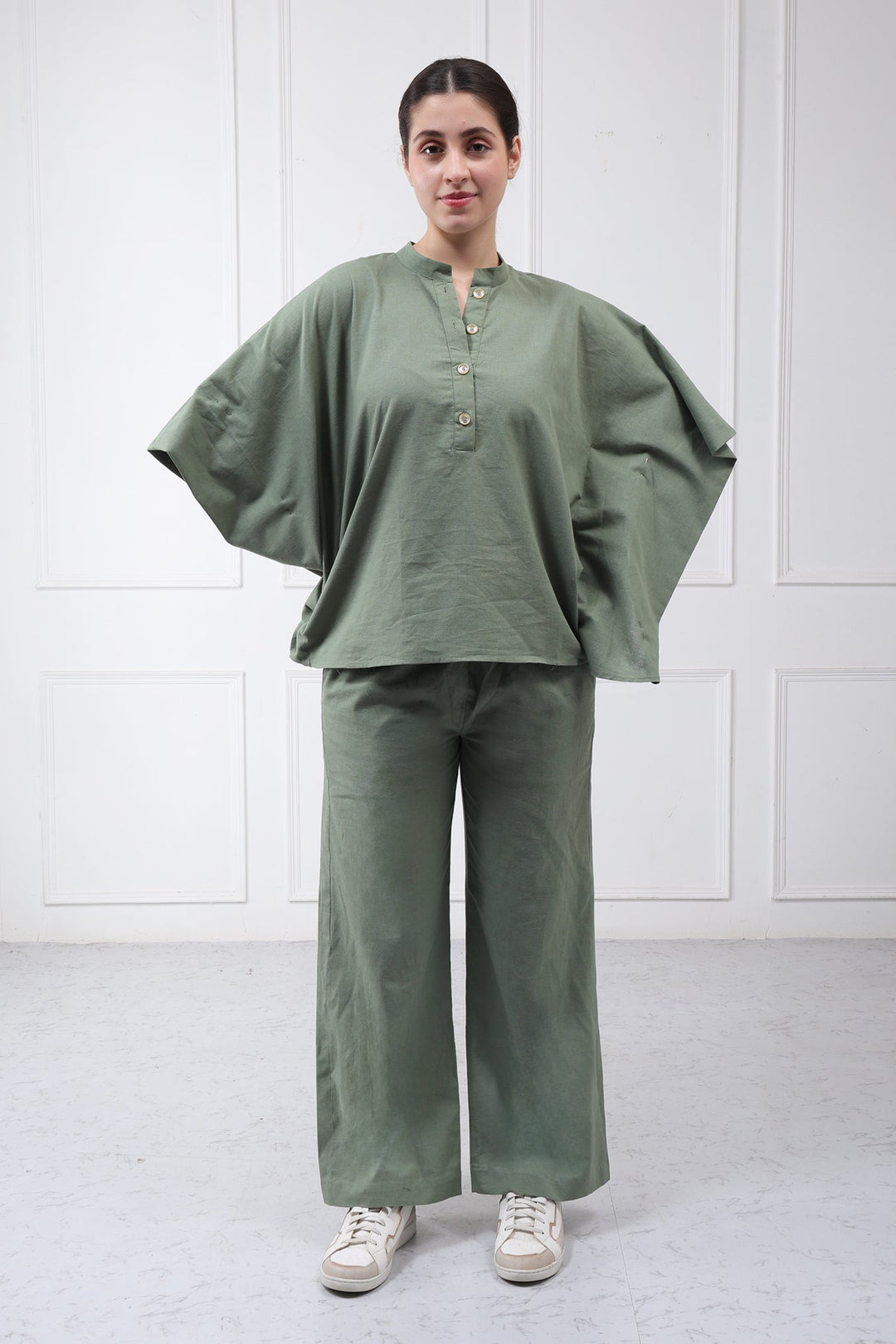 A woman modeling a matching sage green two-piece set consisting of a loose, short-sleeved button-up top and wide-leg trousers, paired with white sneakers, set against a white paneled wall.