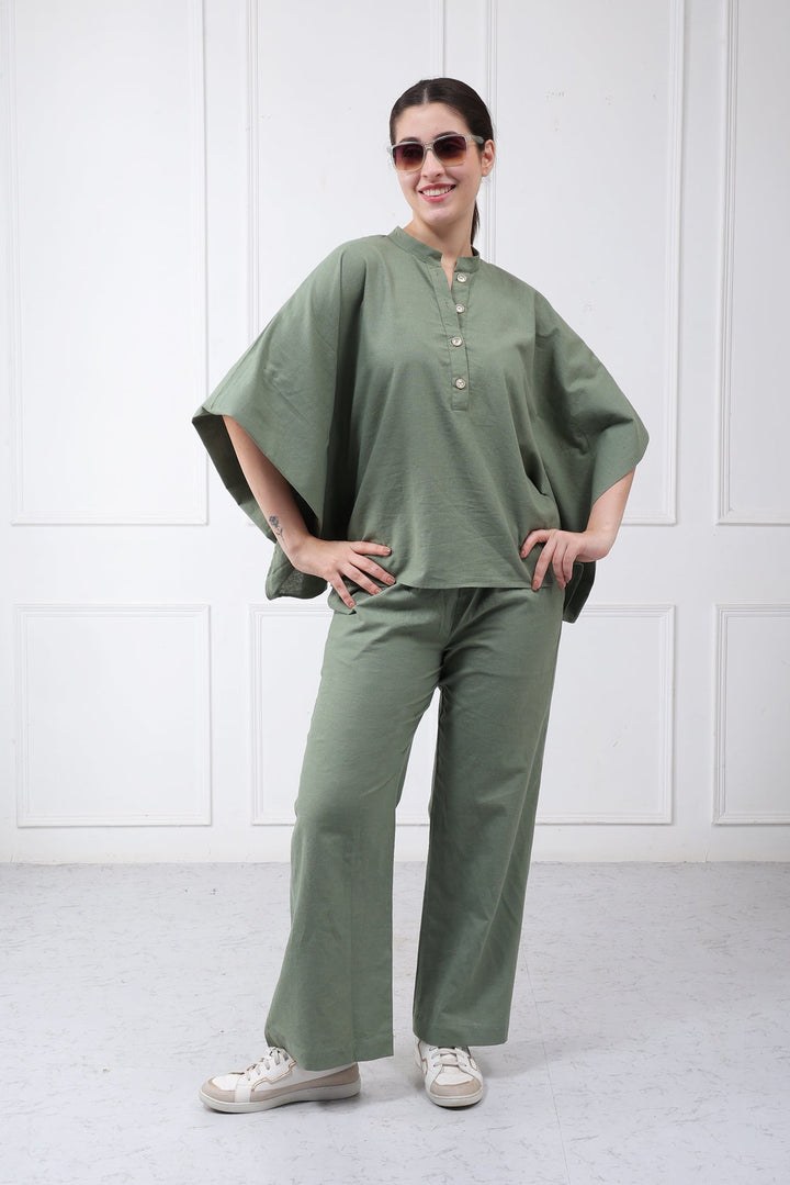 A woman wearing a sage green two-piece linen set, consisting of a loose, wide-sleeved top and matching wide-leg trousers, shown from a rear three-quarter view against a white paneled wall.