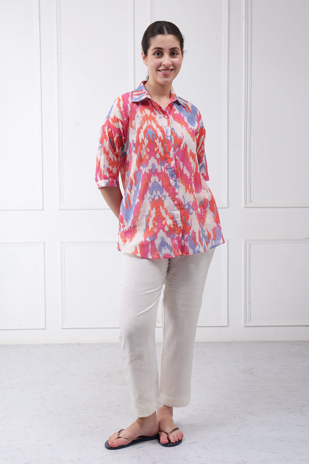 A woman modeling a short-sleeve collared button-up shirt with a vibrant pink, red, and blue abstract ikat pattern, paired with beige trousers and black sandals, standing against a white paneled wall.