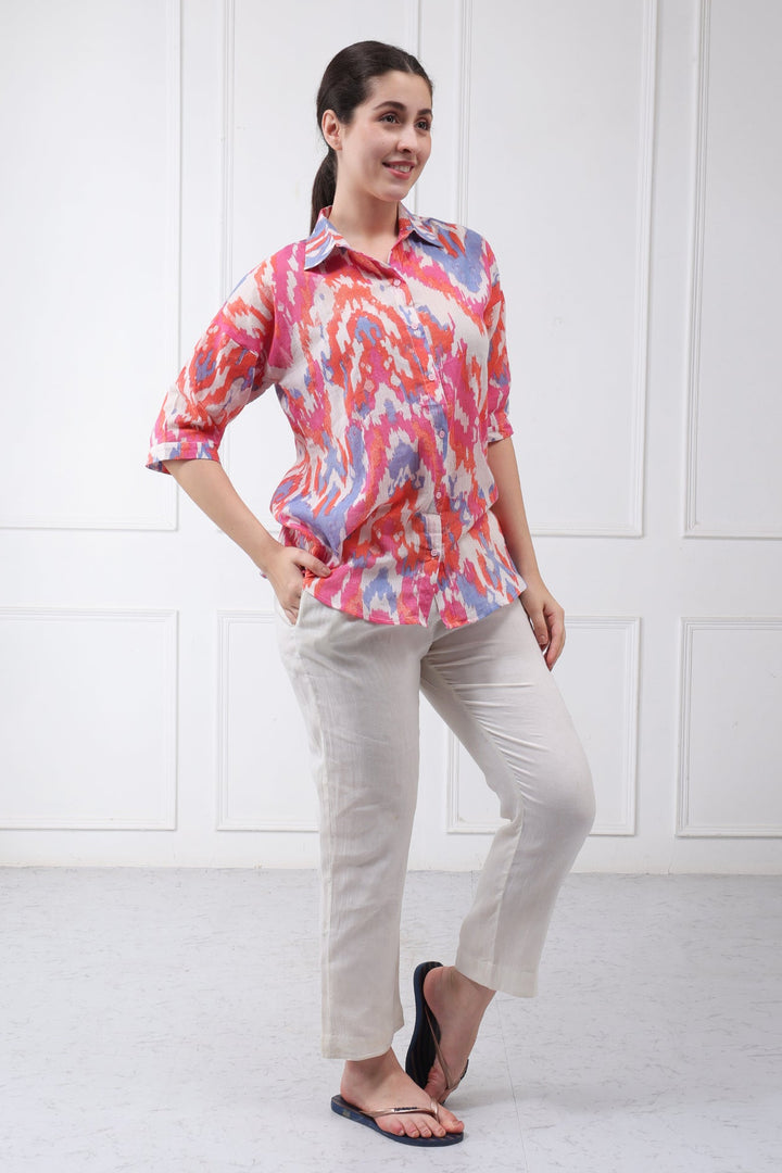 A woman modeling a short-sleeve collared button-up shirt with a vibrant pink, red, and blue abstract pattern, paired with light beige trousers and black sandals, standing against a white paneled wall.