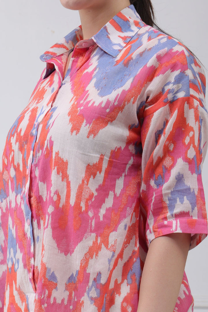 A close-up of a woman wearing a short-sleeve button-up shirt with a vibrant pink, orange, and light blue ikat pattern on a white base.