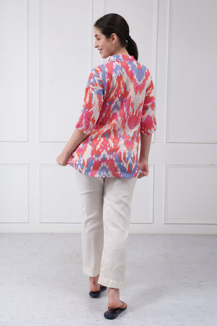 A woman is shown from the back wearing a colorful patterned button-up shirt with three-quarter sleeves, cream trousers, and black sandals, standing against a white paneled wall.