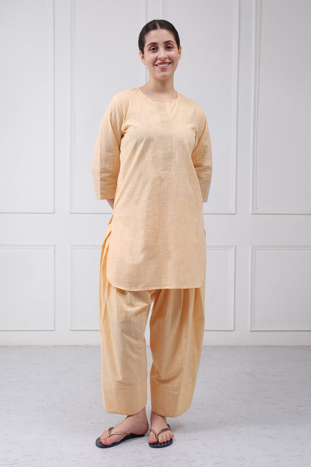 A woman modeling a light peach-beige three-piece kurta set with wide-leg pants and a matching tunic, standing against a white paneled wall.